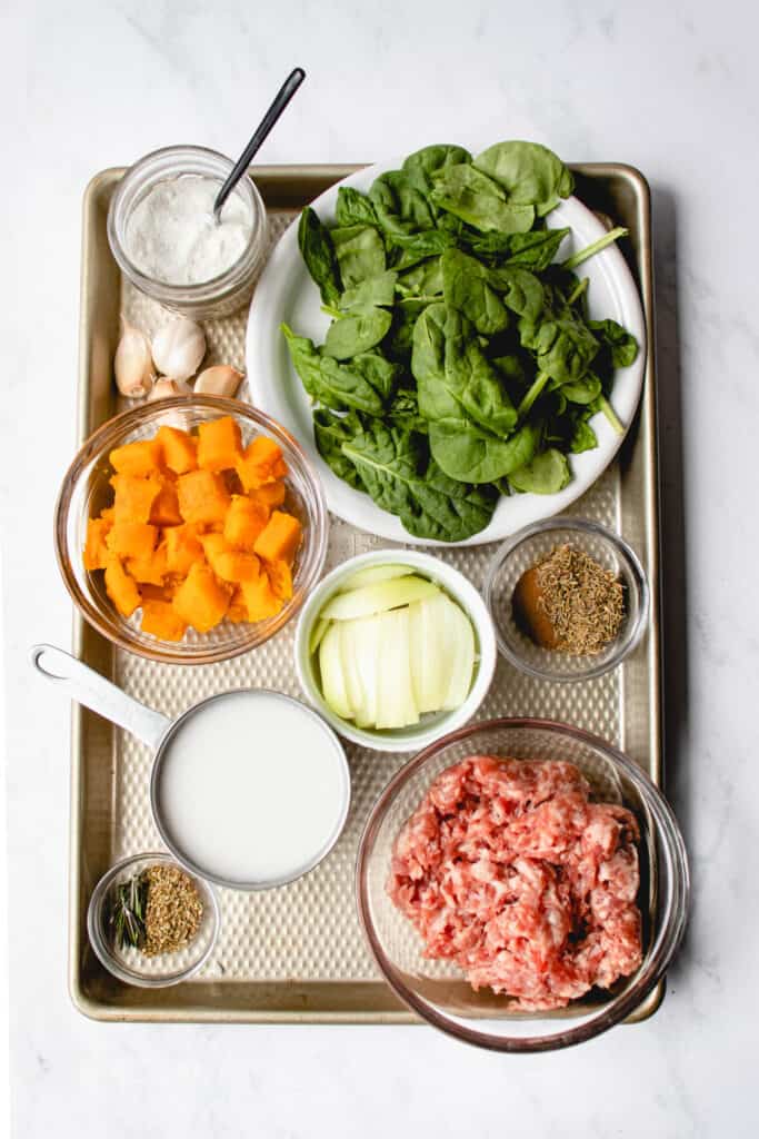 stainless steel sheet pan with a small glass jar of salt with a black spoon, 3 cloves of garlic, glass bowl of cubed butternut squash, white plate with spinach leaves, small white bowl with sliced onion, glass bowl with cinnamon and thyme, stainless steel measuring cup with coconut milk, class bowl with ground pork and small glass bowl with oregano and rosemary