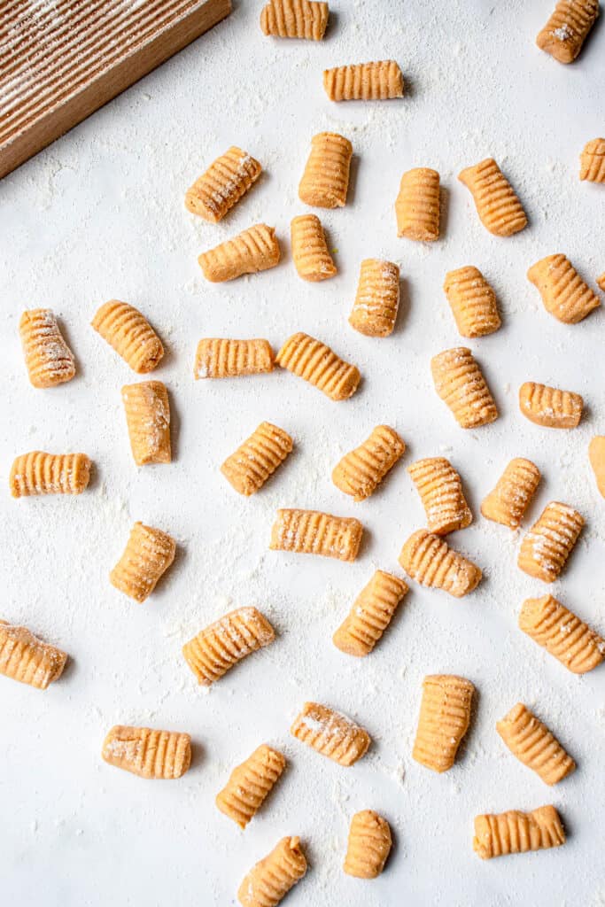 uncooked sweet potato gnocchi pieces on a white marble countertop sprinkled with flour with a wooden gnocchi board in the top left of the image