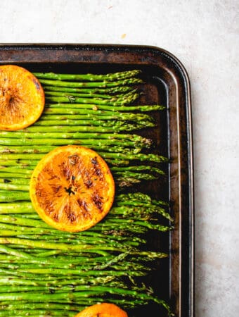 brown sheet pan with grilled asparagus spears topped with sliced, grilled oranges on a light brown background