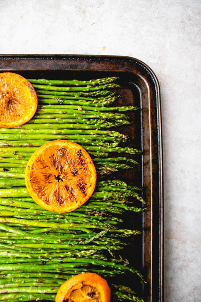 brown sheet pan with grilled asparagus spears topped with sliced, grilled oranges on a light brown background 