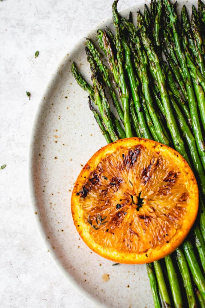 light brown speckled plate with grilled asparagus and one slice of an orange that is grilled with some thyme leaves scattered around the plate