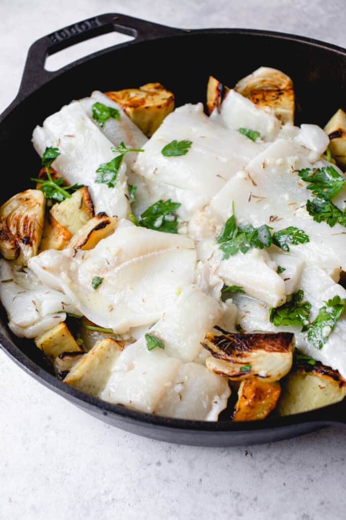 black cast iron pan on a white backdrop filled with roasted sweet potatoes, caramelized fennel and fresh parsley and raw white fish laid on top garnished with fresh parsley ready to be baked