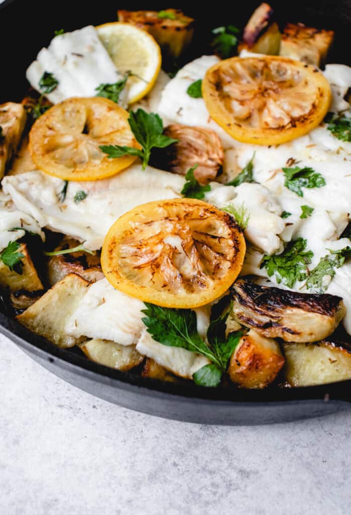 black cast iron pan on a white backdrop filled with baked lemon, white fish, roasted sweet potatoes, caramelized fennel and fresh parsley sprinkled on top