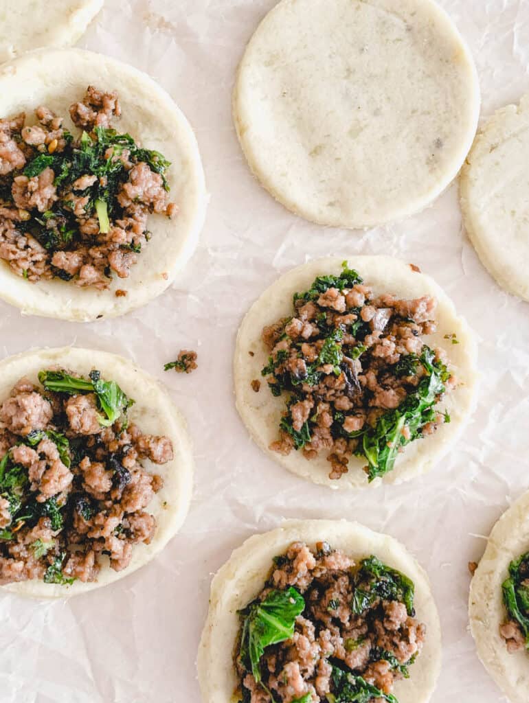 sweet potato and cassava flour circles of dough with ground beef and kale mixture placed within the center of five of the seven circles of dough on a piece of parchment paper