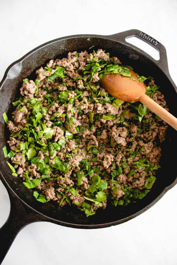 black cast iron skillet with ground beef, kale, parsley and a wooden spoon against a white backdrop