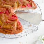 rhubarb upside down cake on a stainless steel cooling rack with 1-inch pieces of rhubarb arranged diagonally on on the top, with a large slice cut out revealing the stainless steel serving knife. there is a white plate with one slice of the cake in the top right of the frame and in the bottom right of the frame there is the head of the knife with some rhubarb smeared on it