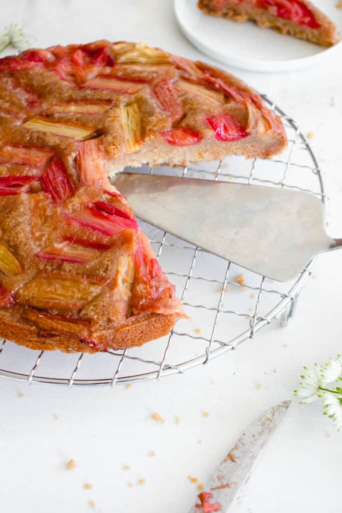 rhubarb upside down cake on a stainless steel cooling rack with 1-inch pieces of rhubarb arranged diagonally on on the top, with a large slice cut out revealing the stainless steel serving knife. there is a white plate with one slice of the cake in the top right of the frame and in the bottom right of the frame there is the head of the knife with some rhubarb smeared on it