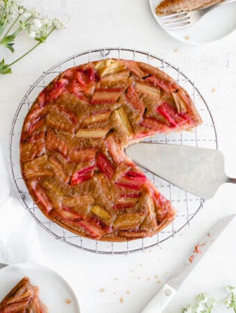 rhubarb upside down cake on a stainless steel cooling rack with 1-inch pieces of rhubarb arranged diagonally on on the top, with a large slice cut out revealing the stainless steel serving knife. the head of a knife with some rhubarb on it is visibile in the bottom right of the frame along with some white flowers. In the bottom left of the image is a white cloth napkin and in the top left of the image are some more white flowers