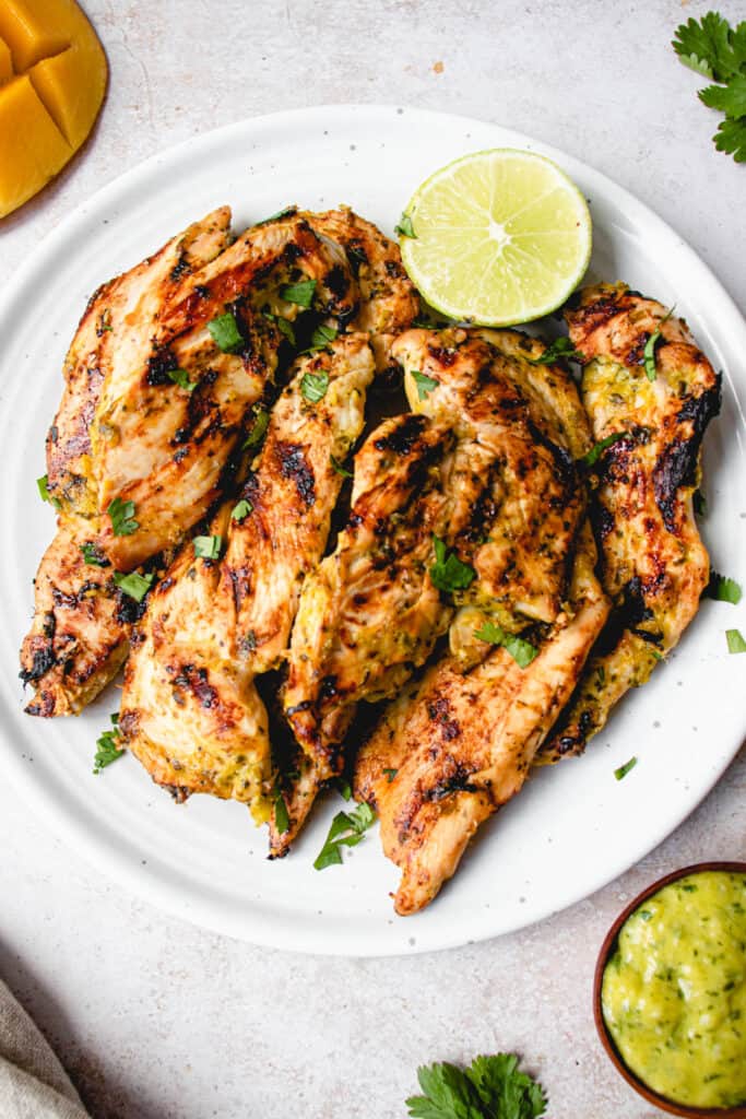 white plate with grilled chicken breast strops garnished with cilantro and a half of a lime on a light grey textured background. in the top left of the frame is a half of a mango scored and in the bottom right of the frame is a small wooden bowl with lime and mango sauce and some cilantro and in the bottom left of the frame there is a light beige cloth napkin