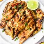 white plate with grilled chicken breast strips garnished with cilantro and a half of a lime on a light grey textured background. in the top left of the frame is a half of a mango scored and in the bottom right of the frame is a small wooden bowl with lime and mango sauce and some cilantro