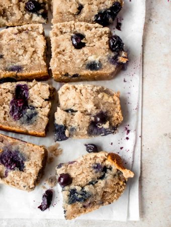 four slices of lemon blueberry banana bread cut into eight squares on two pieces of parchment paper laid on a light grey background