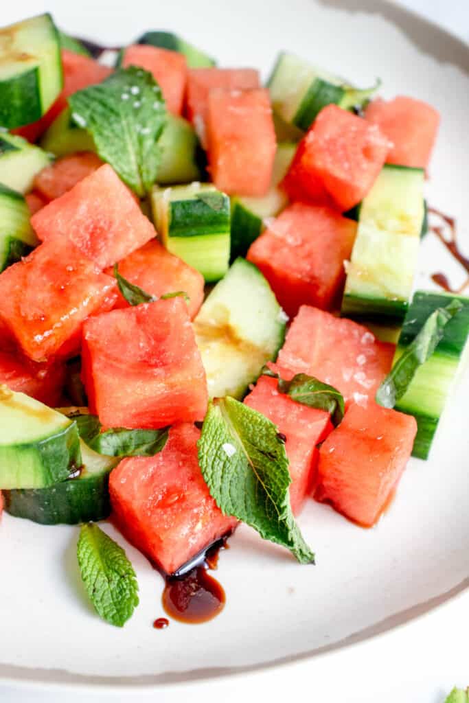 white plate with brown border with cubed watermelon, chopped cucumber, fresh mint, basil, sea salt and balsamic vinegar reduction