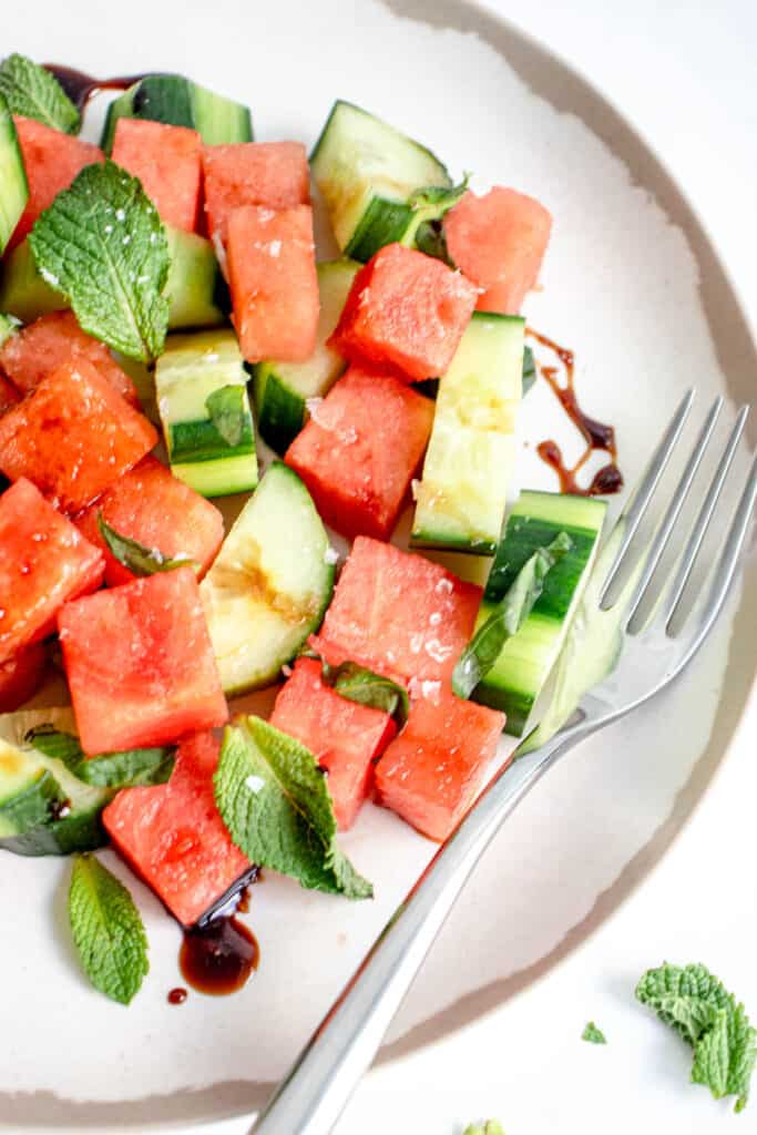 overhead shot of white plate with brown border with cubed watermelon, chopped cucumber, fresh mint, basil, sea salt and balsamic vinegar reduction. There is a stainless steel fork placed on the plate on the right hand side and torn mint leaves to the right of the plate against a white background