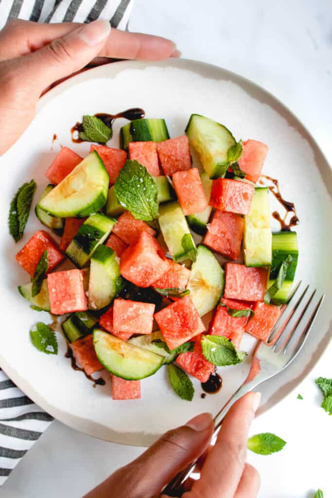 overhead shot of white plate with brown border with cubed watermelon, chopped cucumber, fresh mint, basil, sea salt and balsamic vinegar reduction, there are two brown hands surrounding the plate with one hand placing a stainless steel fork onto the right hand side of the plate. there are torn mint leaves to the right of the plate and a grey and white striped cloth napkin to the left of the plate
