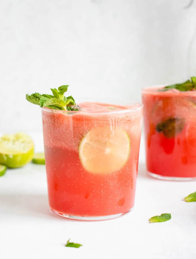Two glasses featuring watermelon and lime juice with sparkling water, fresh mint leaves, a slice of lime and stem of mint leaves garnishing the glass in the front of the frame. There are mint leaves scattered against a white tabletop and there is a lime cut into slices in the background