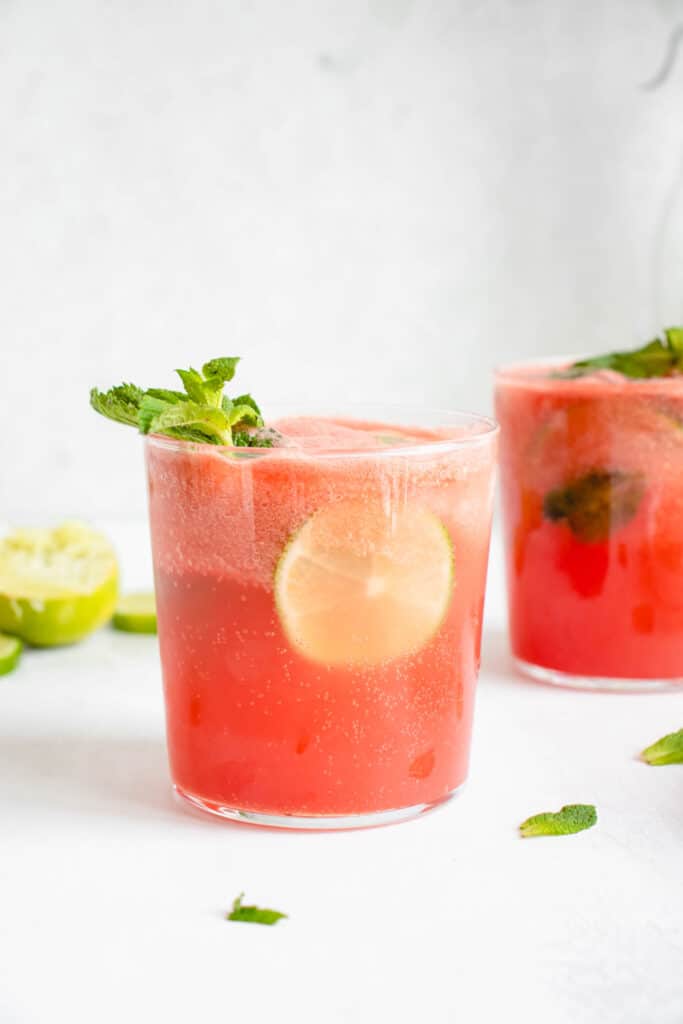Two glasses featuring watermelon and lime juice with sparkling water, fresh mint leaves, a slice of lime and stem of mint leaves garnishing the glass in the front of the frame. There are mint leaves scattered against a white tabletop and there is a lime cut into slices in the background.