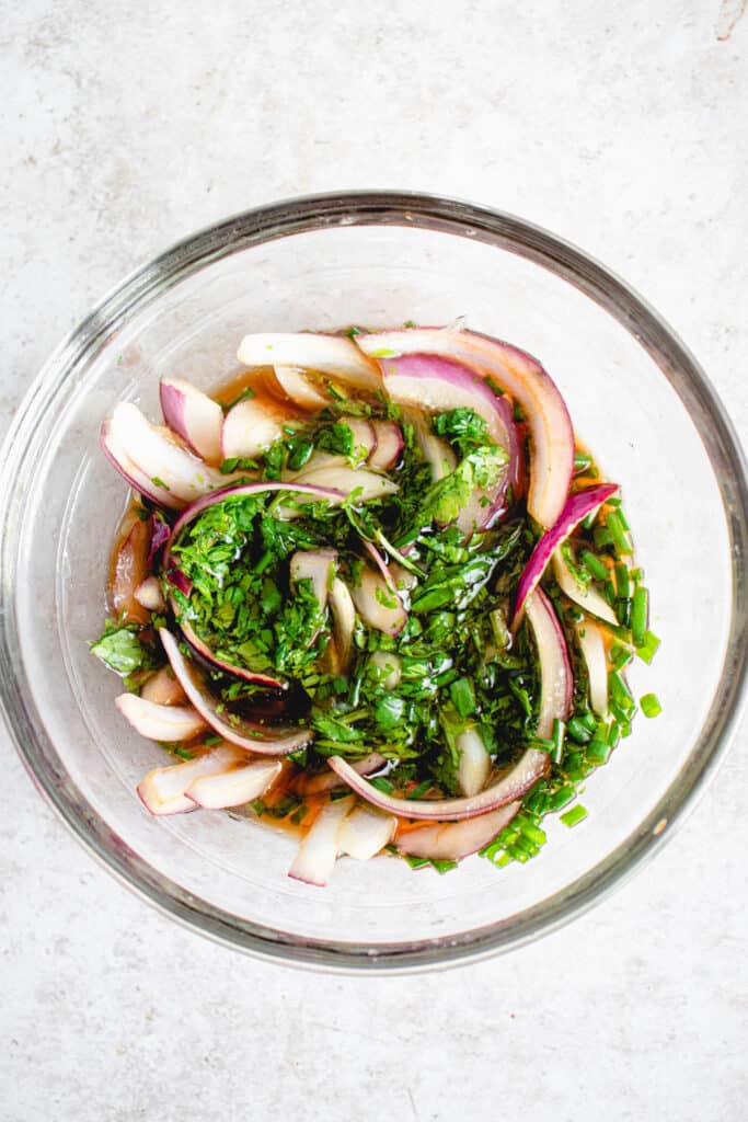 glass bowl with chopped green onions, chopped cilantro and sliced red onion marinating in a lime and coconut aminos dressing on a light grey tabletop