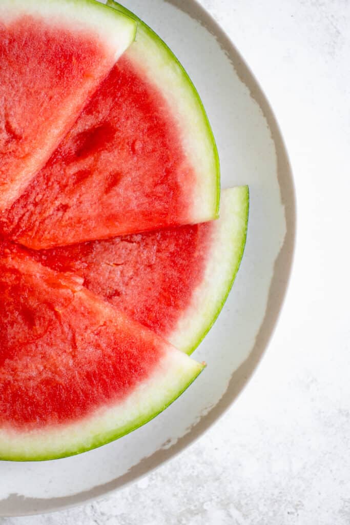 Overhead close up shot of a white plate with brown border with four triangular slices of seedless watermelon. The watermelon slices are laid out in a semi circle. The plate is set against a white background.