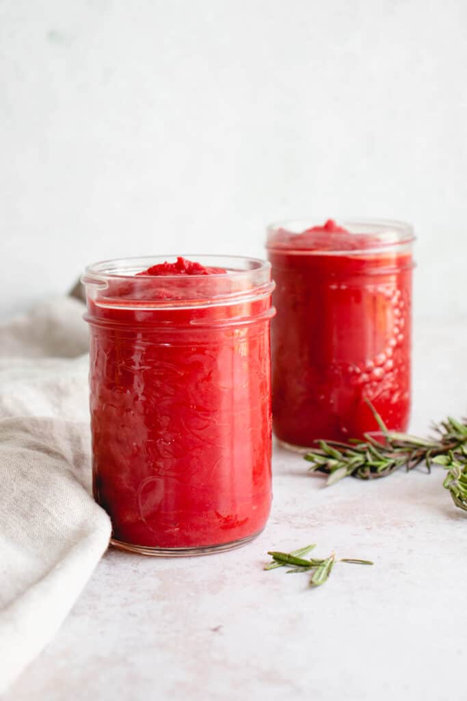 two mason jars filled with nomato sauce diagonally placed on a beige table top with a light beige cloth napkin to the left of the jars there are two sprigs of fresh rosemary placed to the right or the jars and a few leaves of rosemary scattered toward the front of the frame