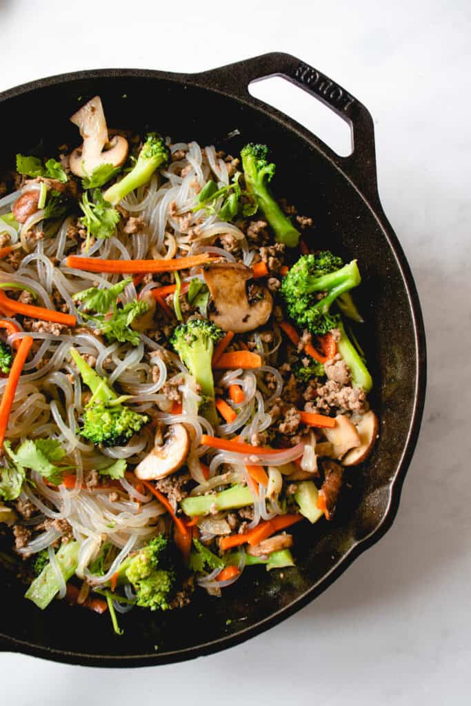 overhead shot of a black cast iron skillet with ground beef, sweet potato noodles, broccoli, julienned carrots and sliced mushrooms. The skillet is on a white backdrop.