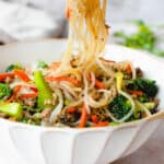 photograph of brown, wooden chopsticks lifting sweet potato noodles out of a white bowl with a brown border. The bowl contains a stir fry of sweet potato noodles, julienned carrots, broccoli florets, sliced mushrooms and ground beef. The bowl is on a grey table top. Unfocused in the background, there is a grey napkin, blurred, in the top left corner. in the top right corner, there are some stems of cilantro, blurred.