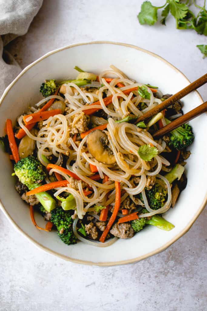 overhead shot of a white bowl with light beige border. in the bowl is a stir fry of sweet potato noodles, broccoli, carrots, mushrooms and ground beef. Two brown wooden chopsticks lay on the top right corner of the bowl. The bowl is on a light grey tabletop. In the top left corner, there is a light grey linen napkin. in the top right corner of 
there are some stems of cilantro.