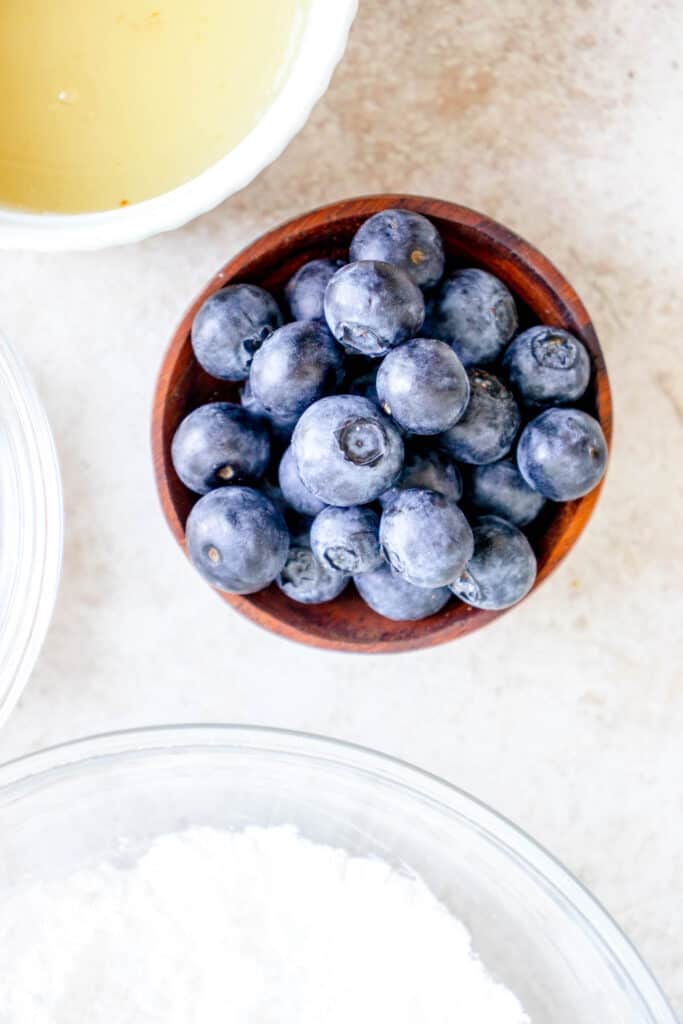 small wooden bowl with blueberries in the center of the image with surrounding clear glass bowls filled with flour, and a white ceramic bowl with gelatin powder, apple cider vinegar and water that is a pale yellow liquid 