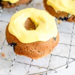 three mini blueberry donuts with a thick yellow glaze arranged on a circular wire cooking rack against a light beige background