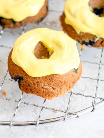 three mini blueberry donuts with a thick yellow glaze arranged on a circular wire cooking rack against a light beige background