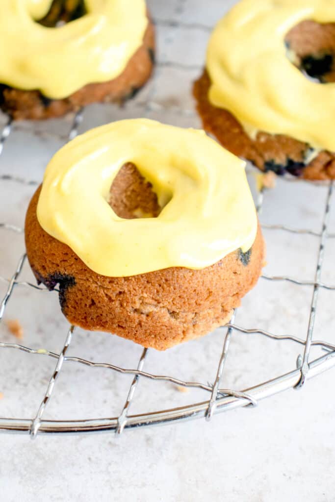 three mini blueberry donuts with a thick yellow glaze arranged on a circular wire cooking rack against a light beige background