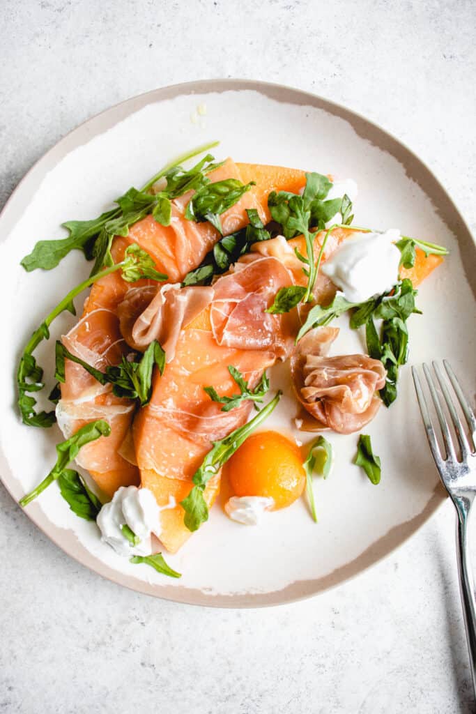 white plate with a brown border with two slices of cantaloupe wrapped in prosciutto, a few arugula laves, one ball of melon and dollops of coconut yogurt drizzled in olive oil. There is a stainless steel fork to the right of the plate which rests on a light grey background 