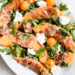 white oval platter with slices of cantaloupe wrapped in prosciutto, a few arugula leaves, balls of melon and dollops of coconut yogurt drizzled in olive oil and sea salt with basil leaves