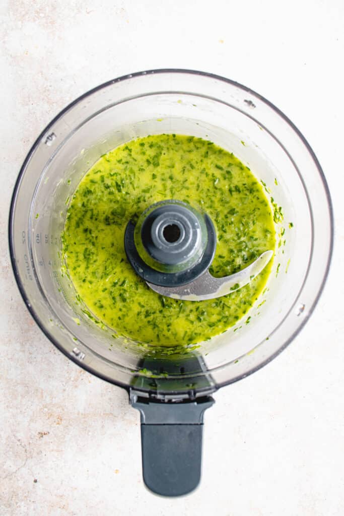 overhead shot of the bowl of a food processor with herbed green marinade against a beige textured background