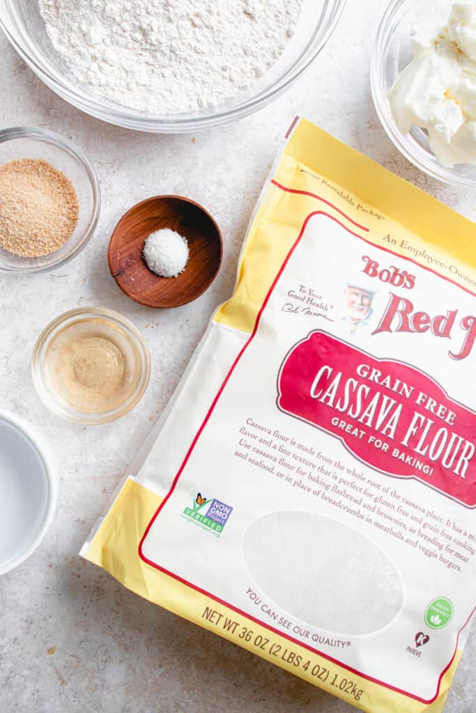 six small glass and wooden bowls with cassava flour, maple sugar, sea salt, apple cider vinegar, water and frozen palm shortening arranged around a yellow, red and white bag of cassava flour against a light beige speckled backdrop