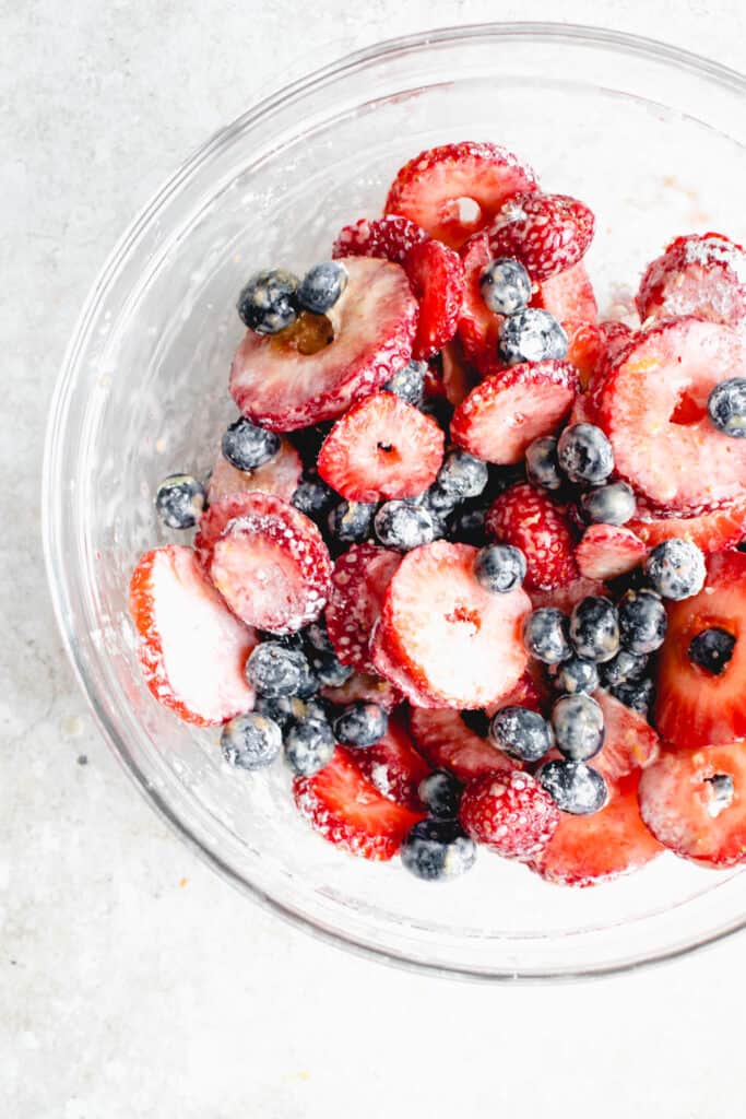 glass bowl with sliced strawberries and blueberries mixed with maple sugar, lemon zest, sea salt, vanilla extract and tapioca starch against a light beige speckled backdrop