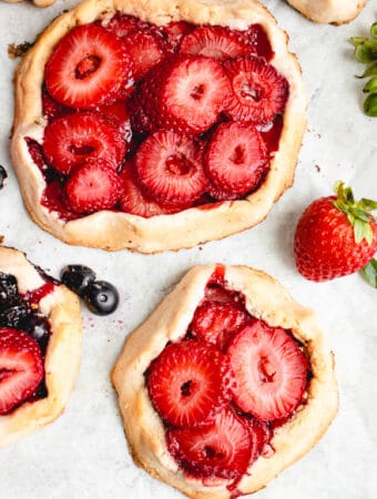 four mini galettes with strawberries and blueberries with a few blueberries and a strawberry scattered around the galettes on a white piece of parchment paper