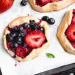three mini galettes with strawberries and blueberries with a few blueberries and a strawberry scattered around the galettes on a white piece of parchment paper inside a brown baking sheet