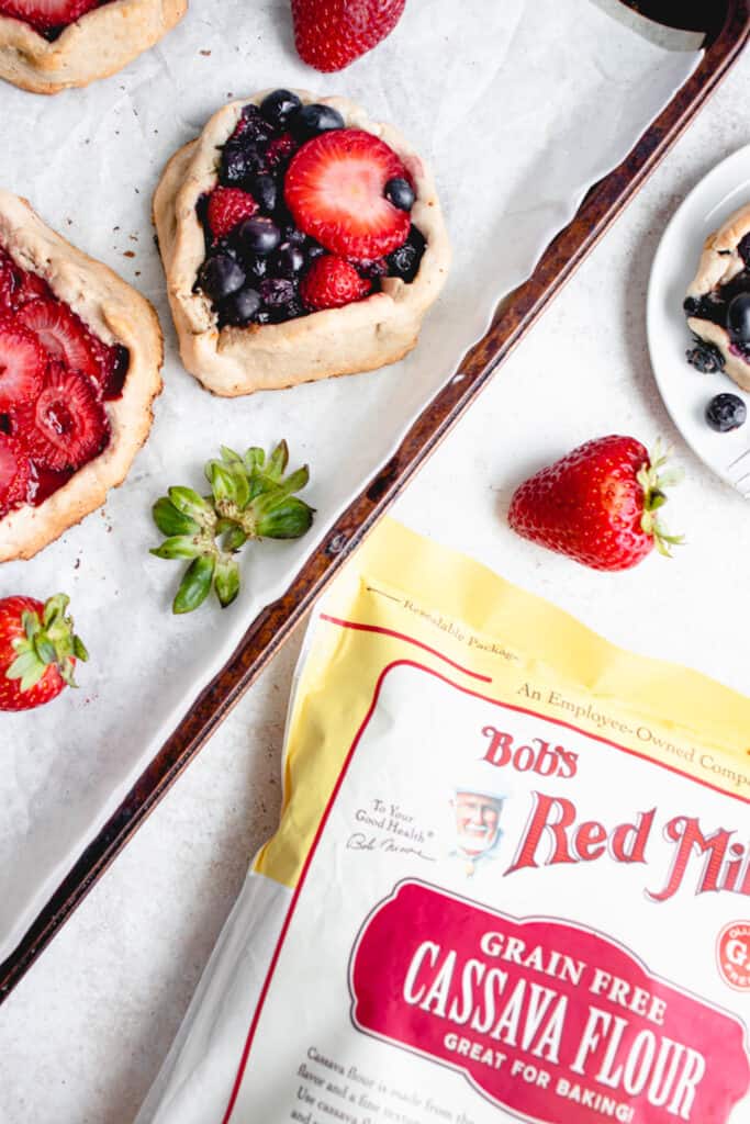 three mini galettes filled with strawberries and blueberries with a few strawberries scattered around the galettes on a white piece of parchment paper inside a brown baking sheet. To the right of the baking sheet which is positioned at an angle there is a small white plate with a blueberry galette and a strawberry to the side and a yellow, red and white bag of Bob's Red Mill Grain-free Cassava Flour