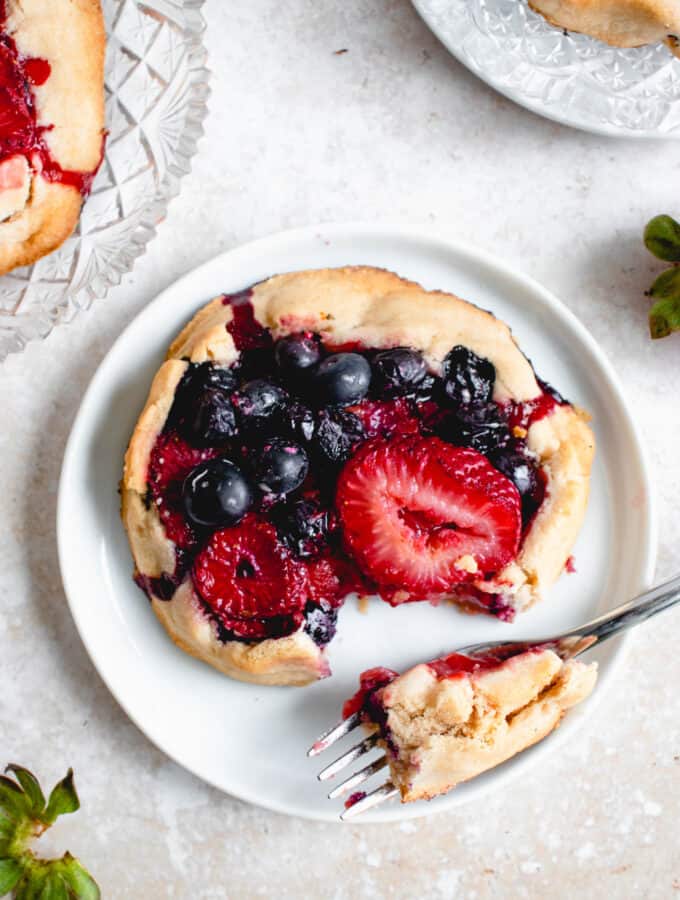 three mini galettes filled with strawberries and blueberries. Two of the galettes are on clear crystal plates and one is on a small white saucer with a piece cut out of it resting on a stainless steel fork