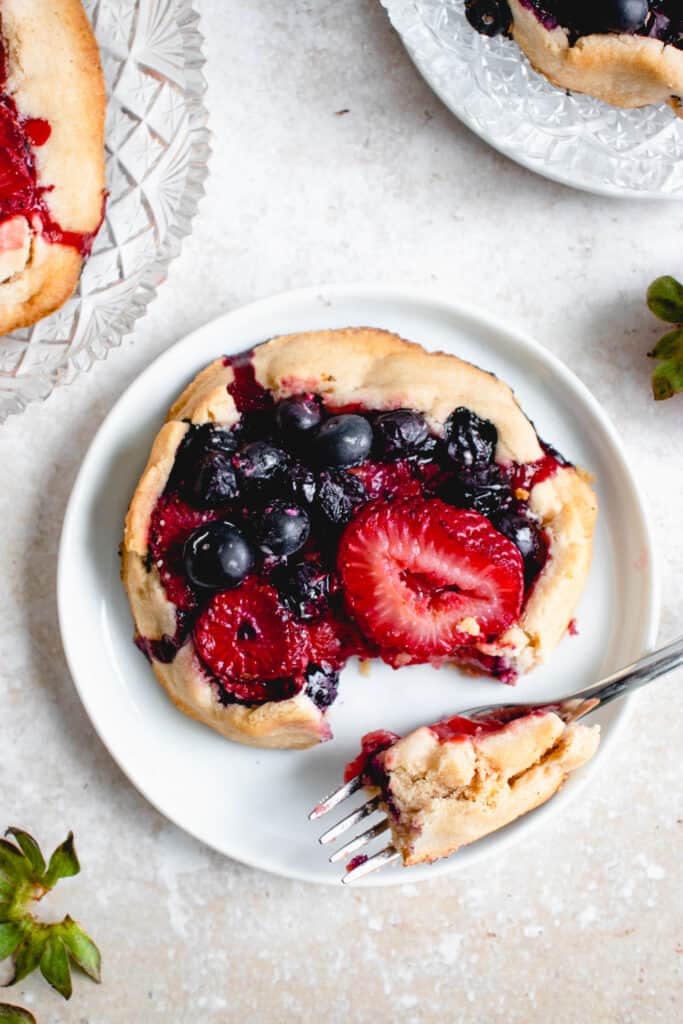 three mini galettes filled with strawberries and blueberries. Two of the galettes are on clear crystal plates and one is on a small white saucer with a piece cut out of it resting on a stainless steel fork