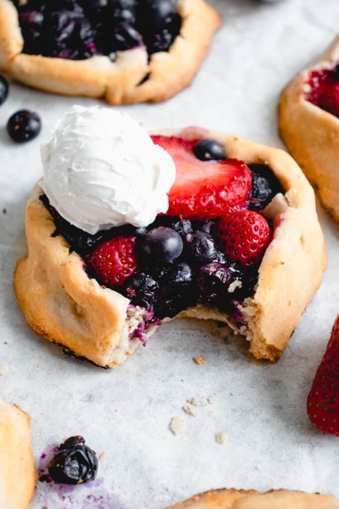 a strawberry blueberry mini galette on a white piece of parchment paper with a bite cut out of the front of the galette and a dollop of whipped coconut cream resting on top. there are three other galettes in the frame with a couple blueberries and a strawberry scattered around the baking sheet