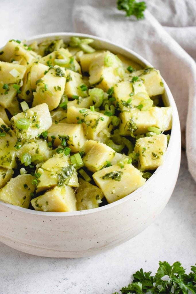 beige bowl with cubed white sweet potatoes, chopped celery, chives, green onions and olive oil against a textured beige and white backdrop. In the background of the image to the right of the bowl is a beige cloth napkin with parsley  