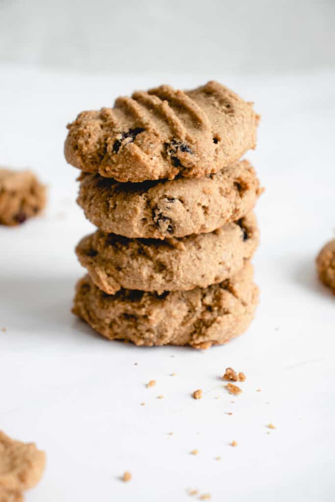 stack of four oatmeal and raisin cookies against a white backdrop with a line of crumbs scattered toward the camera. There are three cookies surrounding the stack of cookies to the back and front left and right. 