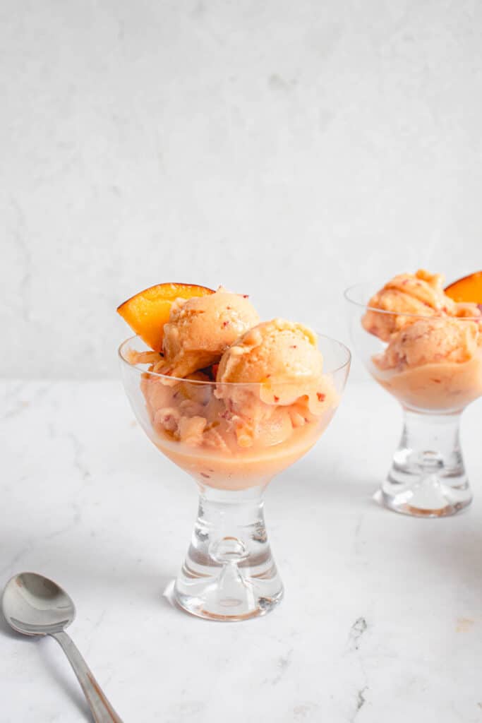 two glass coupes with small scoops of peach ice cream each garnished with a slice of a peach. the glasses are placed diagonally against a grey background with a stainless steel spoon to the left of one coupe