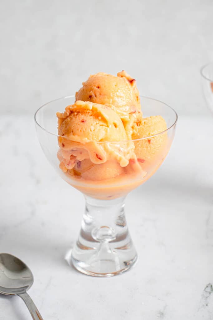 a glass coupe with small scoops of peach ice cream against a grey background with a stainless steel spoon to the left of the coupe