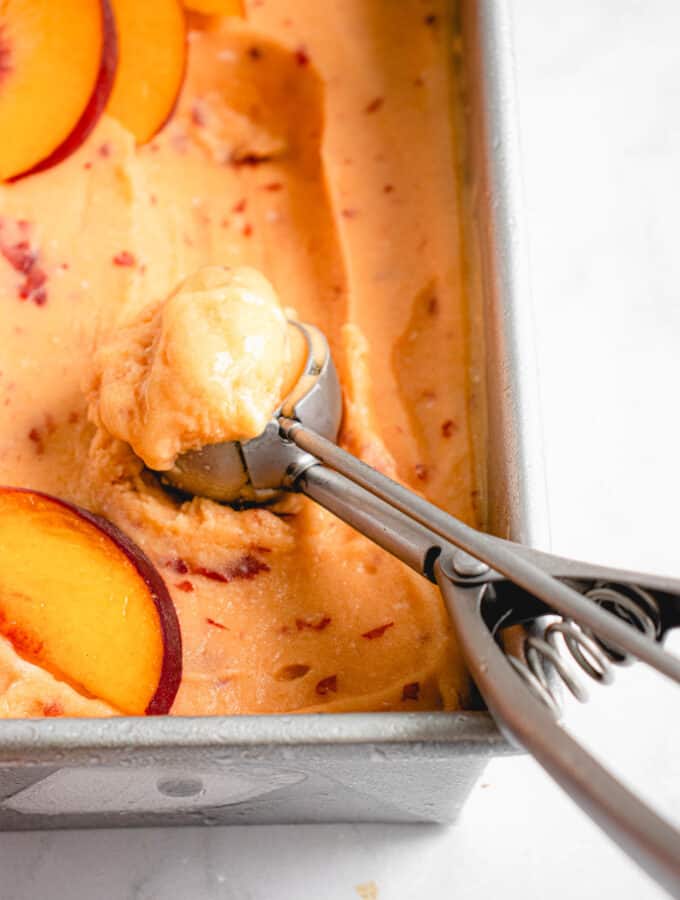 overhead shot of a stainless steel loaf pan with peach ice cream. There is an ice cream scoop filled with a scoop of the ice cream in the bottom left third of the image and there are peach slices laid on top the ice cream