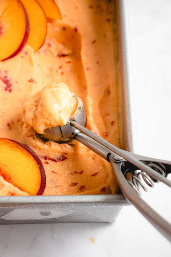overhead shot of a stainless steel loaf pan with peach ice cream. There is an ice cream scoop filled with a scoop of the ice cream in the bottom left third of the image and there are peach slices laid on top the ice cream