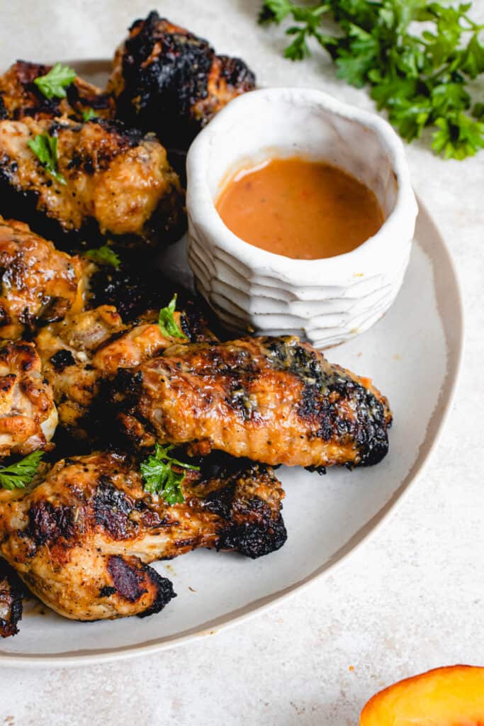 white plate with brown border with chicken wings grilled and glazed with peach-ginger sauce and topped with chopped parsley. to the right of the plate is a small white ceramic bowl with leftover peach-ginger sauce and there are some sprigs of parsley in the back right of the image and a piece of peach in the front right of the image