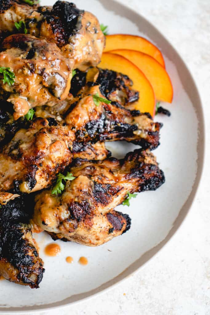 white plate with brown border with chicken wings grilled and glazed with peach-ginger sauce and topped with chopped parsley. There are some sliced peaches tucked under the chicken wings in the top right of the plate and some peach-ginger sauce scattered on top the chicken and onto the white plate