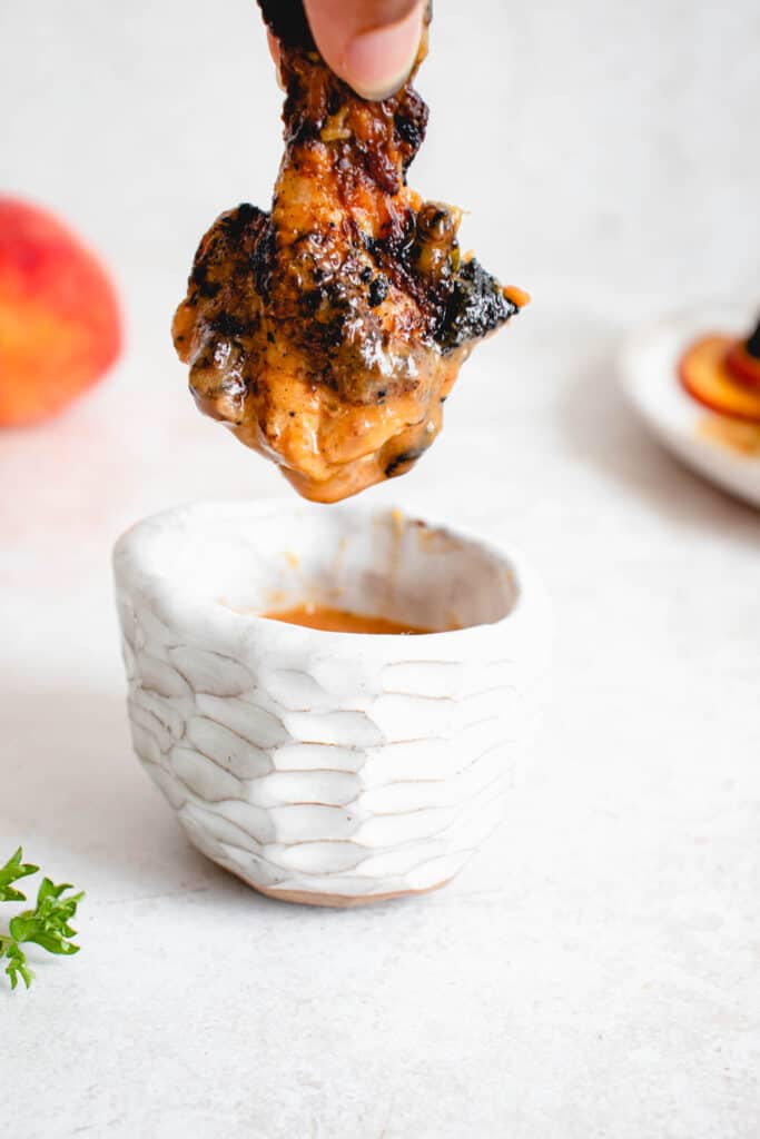  white ceramic bowl with a textured pattern holding peach-ginger sauce with a brown hand holding one small chicken drumstick above the bowl. there are some peaches in the background and a sprig of parsley in the bottom left of the frame